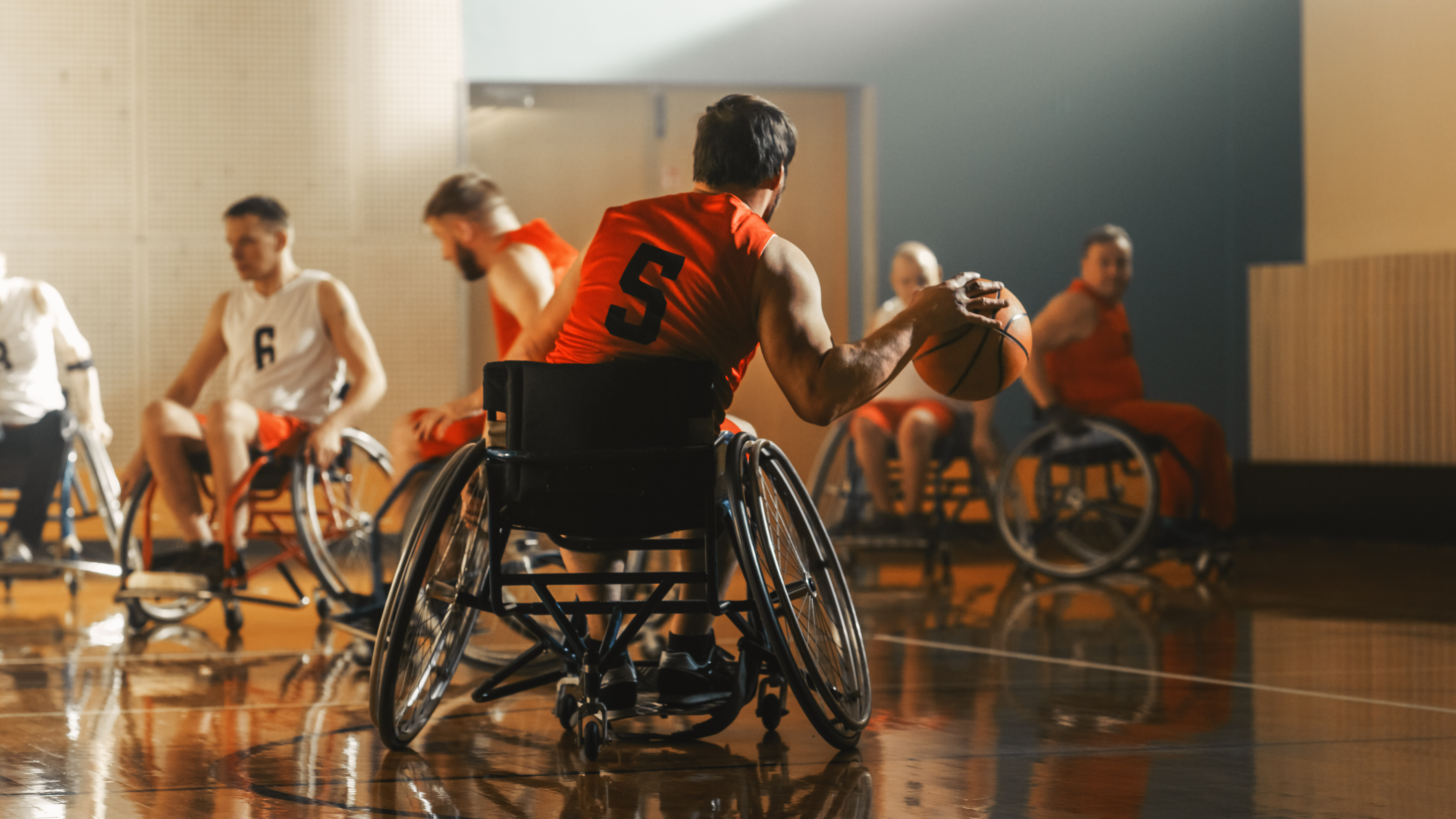 Mehrere Personen im Rollstuhl spielen Rollstuhlbasketball in einer Sporthalle.