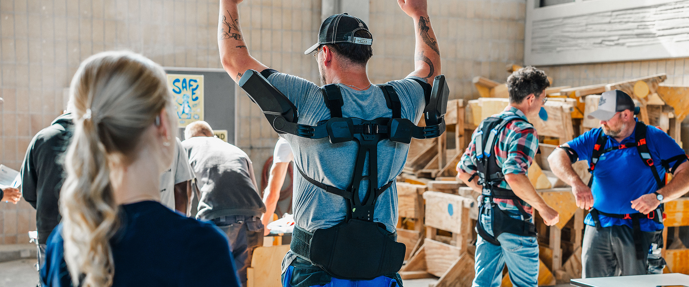 Ein Auszubildender trägt ein Exoskelett und hebt die Arme. Im Hintergrund stehen weitere Menschen mit weiteren Exoskeletten.