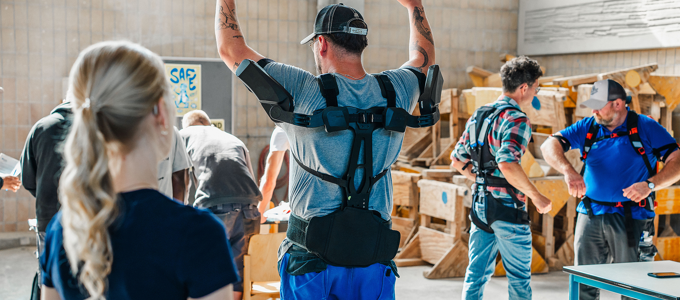 Ein Auszubildender tr&auml;gt ein Exoskelett und hebt die Arme. Im Hintergrund stehen weitere Menschen mit weiteren Exoskeletten.
