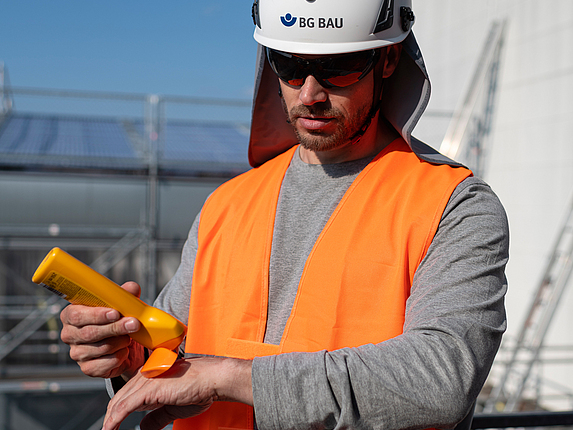 Ein Bauarbeiter cremt sich auf einem Gerüst mit Sonnenschutzcreme ein. Er trägt einen Schutzhelm mit Nackenschutztuch, eine Sonnenbrille sowie eine UV-Weste.