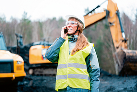 Frau in Warnweste telefoniert auf einer Baustelle mit Bagger.