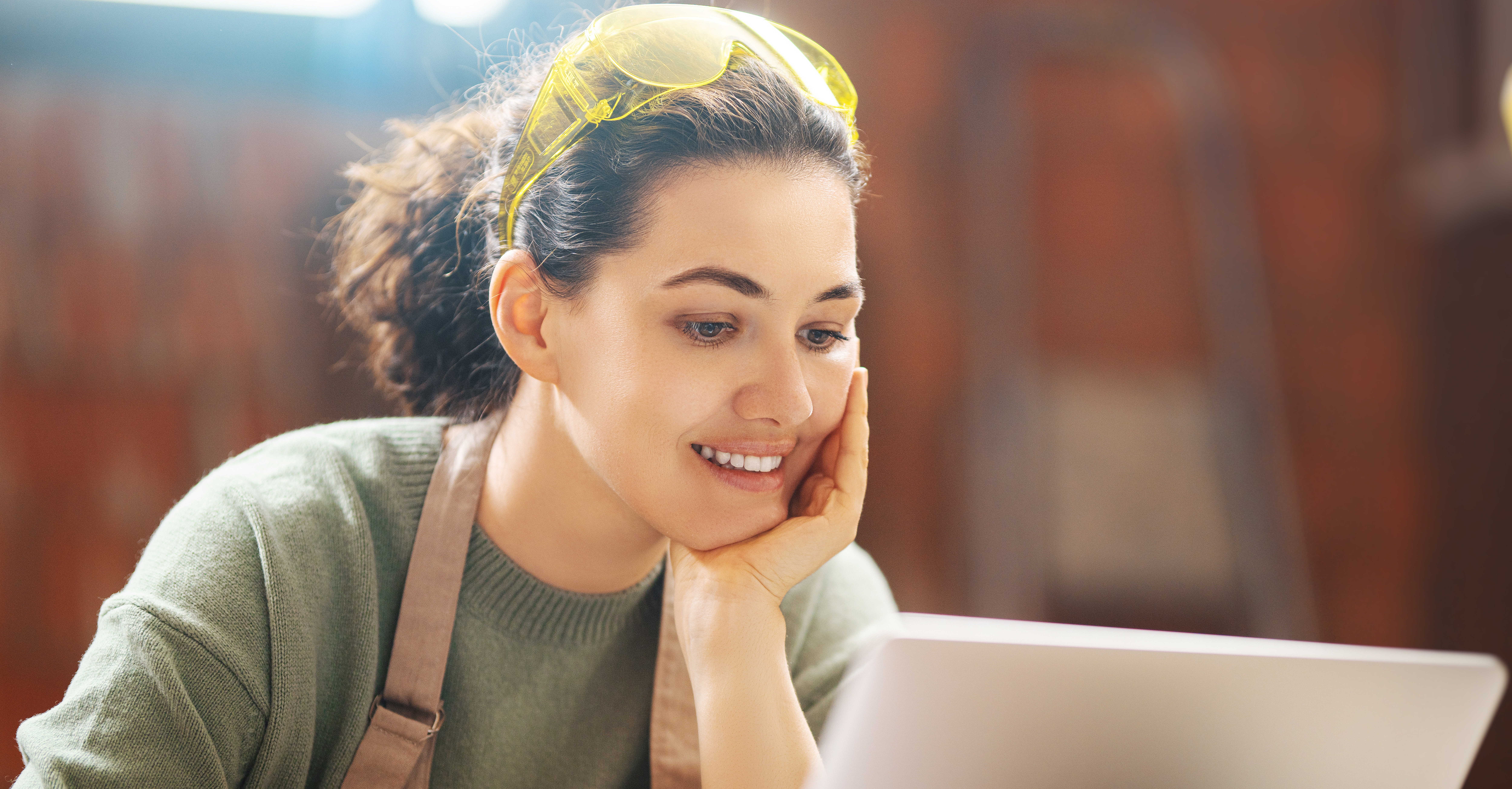 Eine junge Frau sitzt während der Arbeit mit Schutzbrille am Laptop. Sie ist in einem Werkraum.