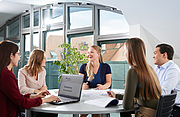 Fünf junge Personen, vier Frauen und ein Mann, sitzen an einem runden Tisch während einer Teamsitzung.