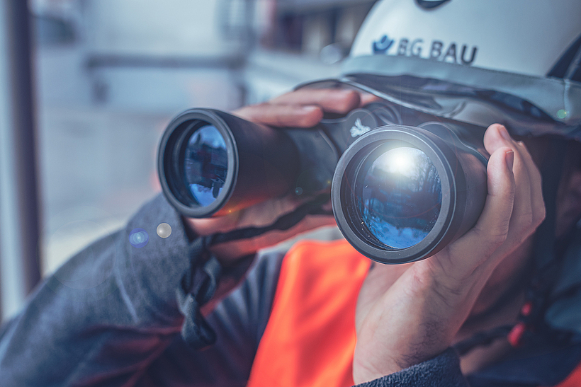 Eine Person mit BG BAU-Schutzhelm blickt durch ein Fernglas.