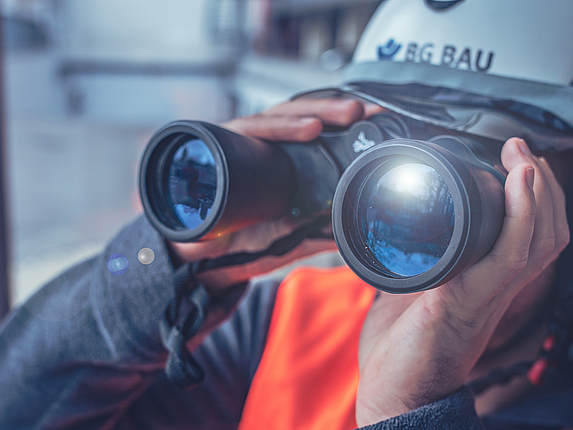 Eine Person mit BG BAU-Schutzhelm blickt durch ein Fernglas.