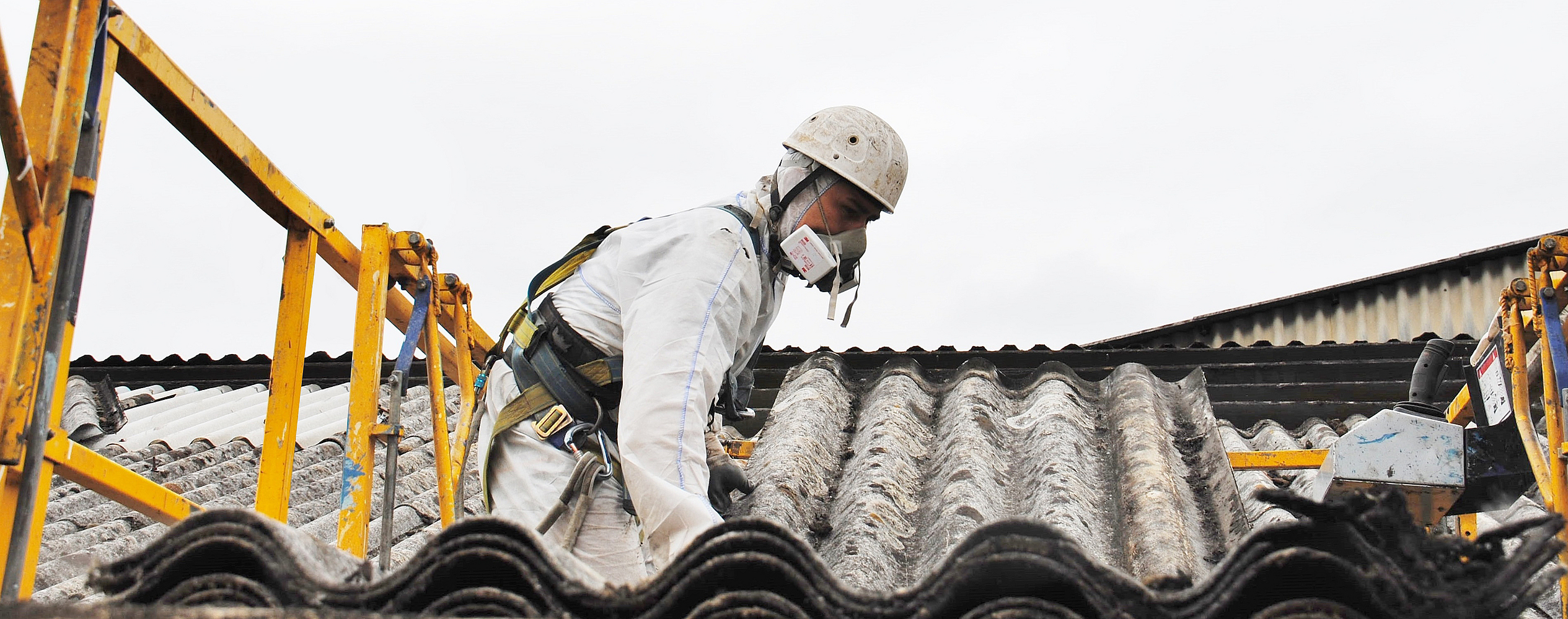 Mann in Schutzanzug mit Atemschutz und Schutzhelm arbeitet auf einem gewellten Dach.