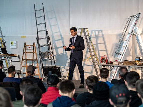 Ein Mann steht auf eine Bühne mit verschiedenen Arten von Leitern und hält einen Vortrag vor einem sitzenden Publikum.
