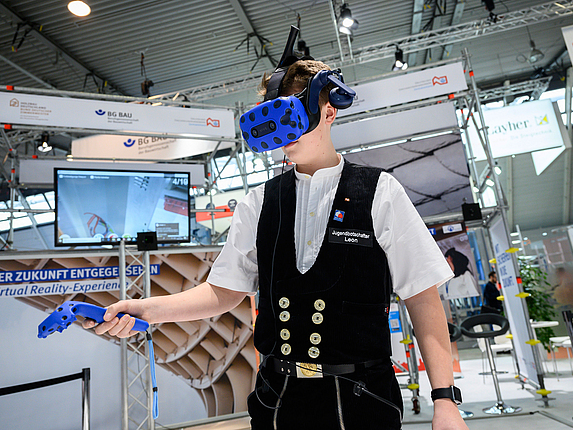 Ein Mann in Zimmermannskleidung trägt an einem Messestand eine VR-Brille und erkundet etwas mit dem Scanner.