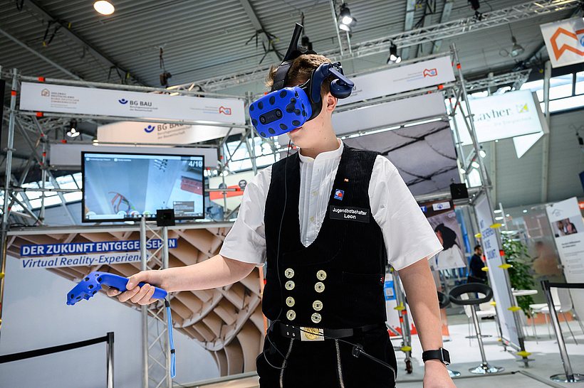 Ein Mann in Zimmermannskleidung trägt an einem Messestand eine VR-Brille und erkundet etwas mit dem Scanner.