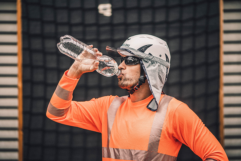 Ein Bauarbeiter mit Schutzhelm, Sonnenbrille und Nackentuch trinkt Wasser in der Pause.