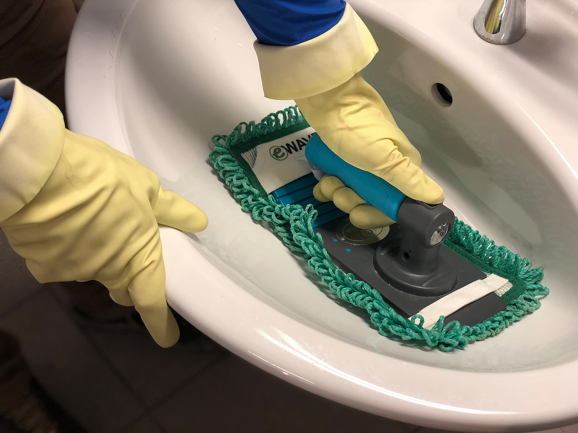 Ein Waschbecken wird mit einem Reinigungsmobb gesäubert. Die Person trägt hellgelbe Schutzhandschuhe.