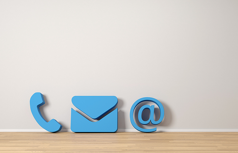 Drei blaue Icons: eine Telefonhörer, ein Brief und @-Zeichen stehen auf einem Holzboden.