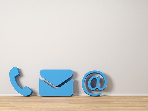 Drei blaue Icons: eine Telefonhörer, ein Brief und @-Zeichen stehen auf einem Holzboden.
