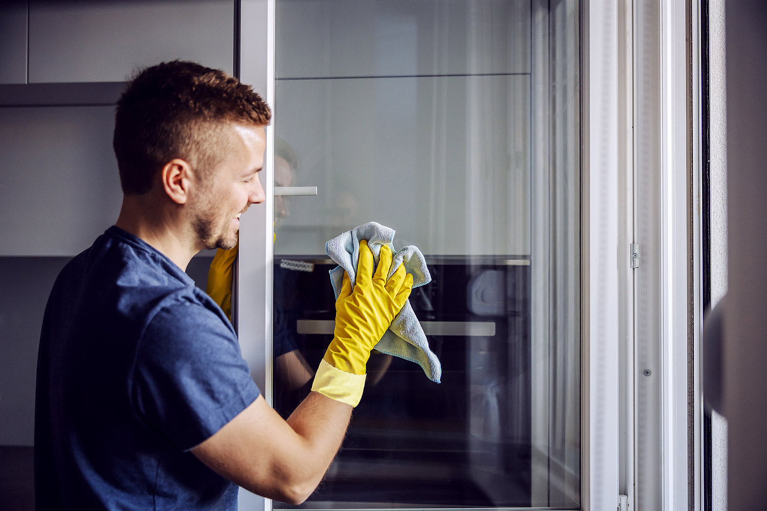 Junger Mann reinigt ein Fenster mit Reinigungstuch und trägt dabei Schutzhandschuhe.