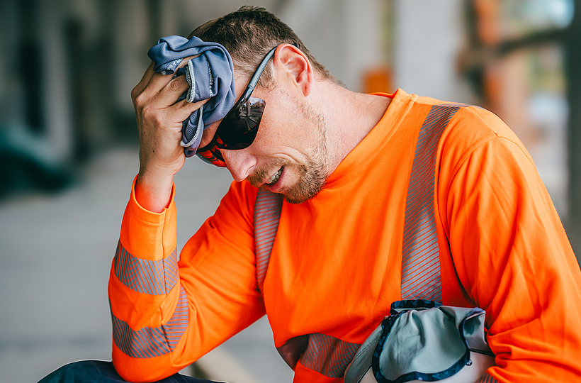 Ein Beschäftigter ruht sich im Schatten von der Arbeit in der Hitze aus. Er trägt dabei ein Funktionsshirt und eine Sonnenbrille. Er kühlt seinen Kopf mit einem Kühltuch und hält den Schutzhelm mit Nackentuch unterm Arm.