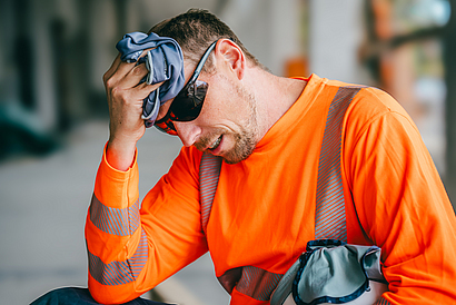 Ein Beschäftigter ruht sich im Schatten von der Arbeit in der Hitze aus. Er trägt dabei ein Funktionsshirt und eine Sonnenbrille. Er kühlt seinen Kopf mit einem Kühltuch und hält den Schutzhelm mit Nackentuch unterm Arm.
