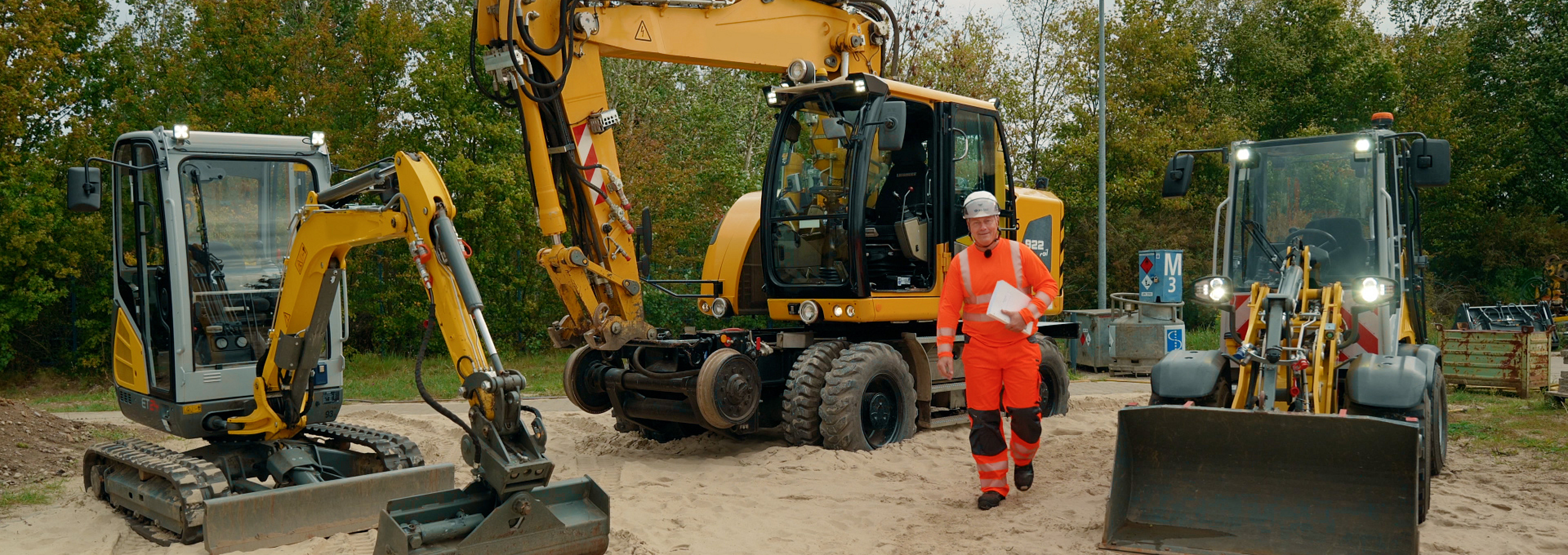 BG BAU Aufsichtsperson Denny Hillert steht neben drei Erdbaumaschinen.