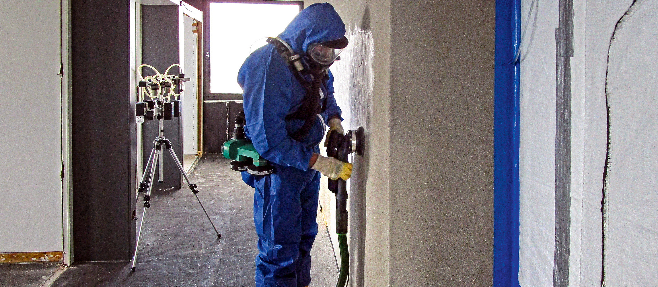 Ein Mann in Schutzanzug und Atemschutzmaske entfernt den Putz von der Wand mit einem Bearbeitungssystem, das mit Absaugung ausgestattet ist.