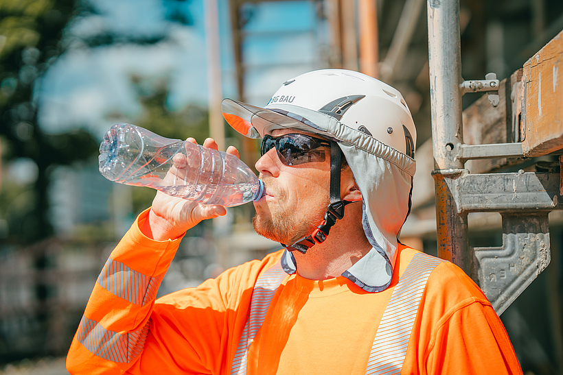 Ein Beschäftigter trinkt Wasser und trägt einen Schutzhelm, eine Sonnenbrille, ein Nackentuch und ein Funktionsshirt. 