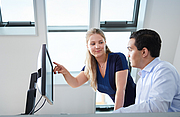 Eine junge Frau zeigt mit ihrem rechten Arm auf den Monitor, während sie ihren Kollegen ansieht.