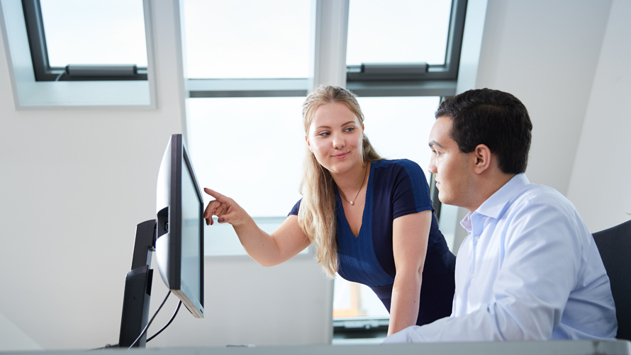 Eine junge Frau zeigt mit ihrem rechten Arm auf den Monitor, während sie ihren Kollegen ansieht.