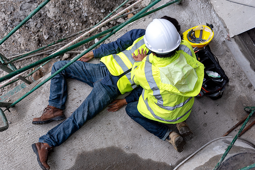 Ein Bauarbeiter hat einen Arbeitsunfall. Ein anderer Bauarbeiter leistet Erste-Hilfe.