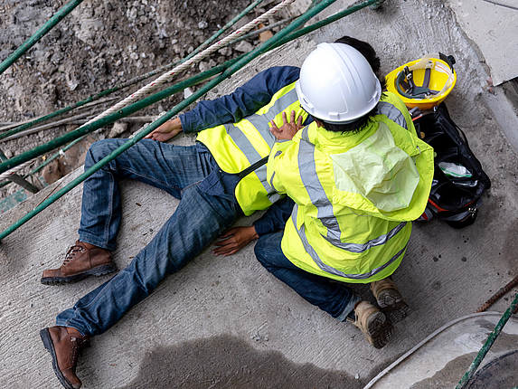 Ein Bauarbeiter hat einen Arbeitsunfall. Ein anderer Bauarbeiter leistet Erste-Hilfe.