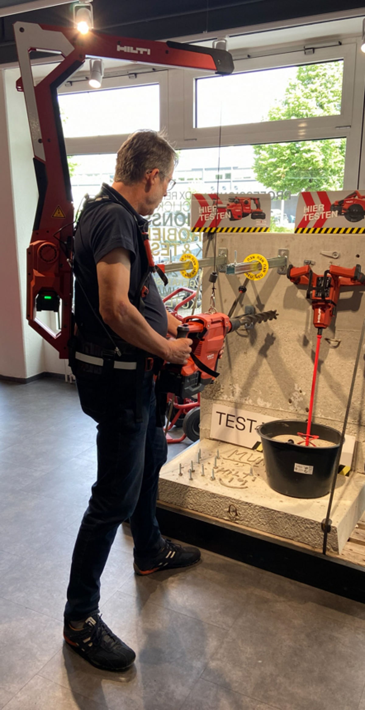 Eine Person trägt einen Balancer am Körper, der das Gewicht eines roten Bohrhammers hält, und arbeitet damit an einer Wand mit ausgestellten Werkzeugen.