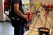 Eine Person trägt einen Balancer am Körper, der das Gewicht eines roten Bohrhammers hält, und arbeitet damit an einer Wand mit ausgestellten Werkzeugen.