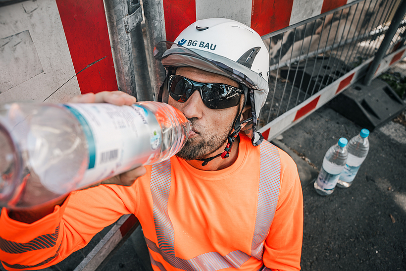 Ein Bauarbeiter in Warnkleidung und Schutzhelm trinkt an einer Baustelle bei heißem Wetter Wasser, um sich abzukühlen.