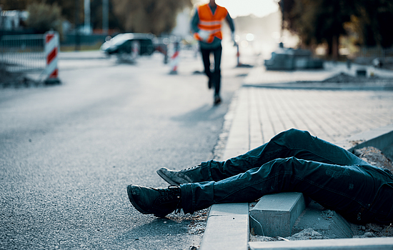 Eine verunglückte Person liegt auf der Straße. Es kommt ein Bauarbeiter zur Hilfe. Im Hintergrund sieht man eine Baustelle.