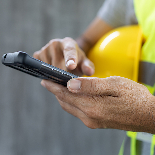 Finger eines Bauarbeiters tippt auf einem Smartphone.