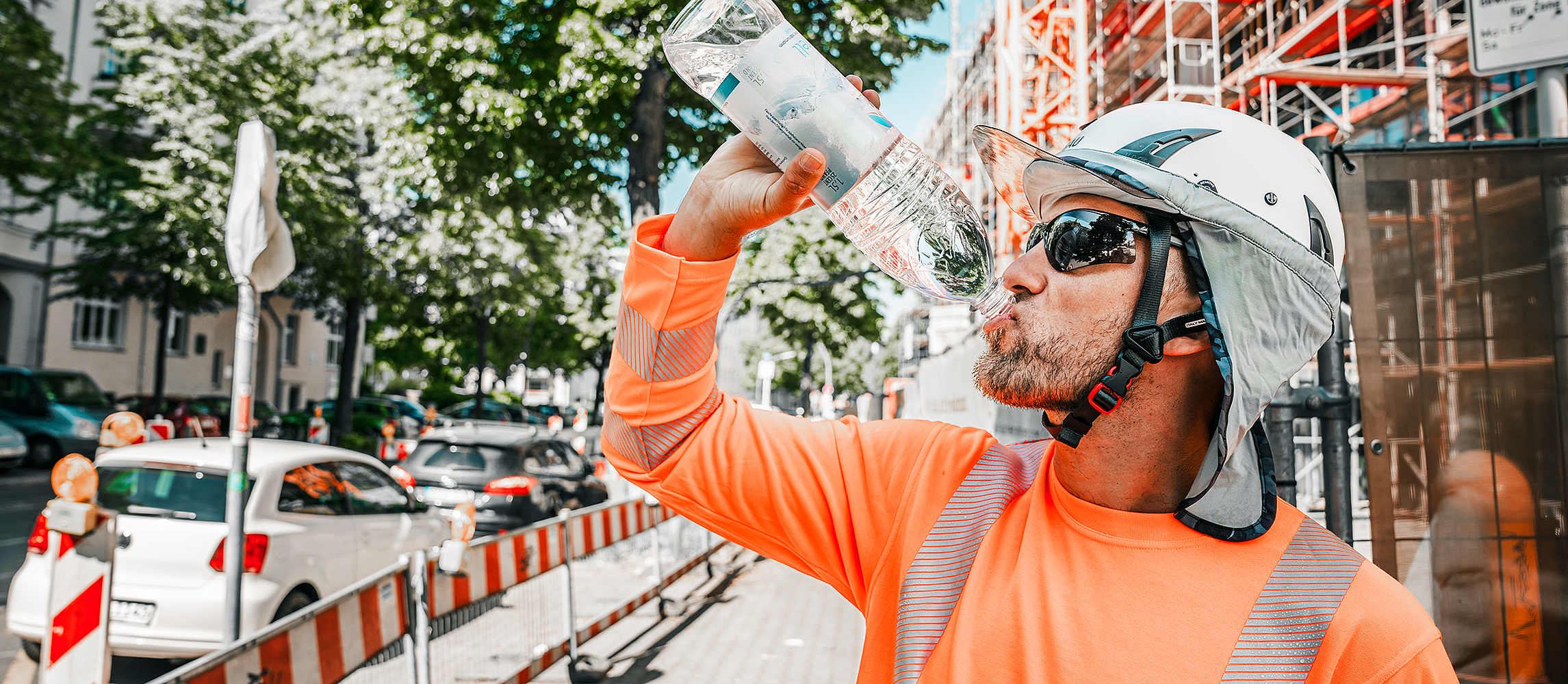 Ein Bauarbeiter in Warnkleidung und Schutzhelm trinkt an einem heißen Tag auf einer städtischen Baustelle Wasser, um sich vor Überhitzung zu schützen.