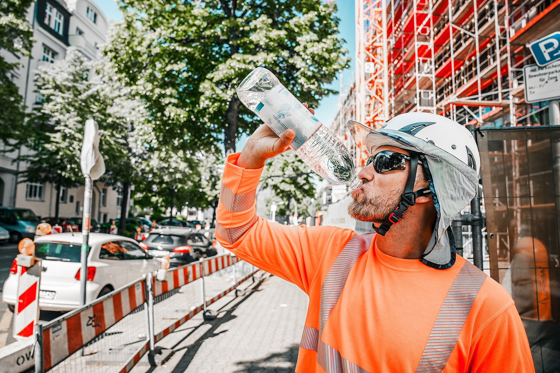 Ein Bauarbeiter in Warnkleidung und Schutzhelm trinkt an einem heißen Tag auf einer städtischen Baustelle Wasser, um sich vor Überhitzung zu schützen.