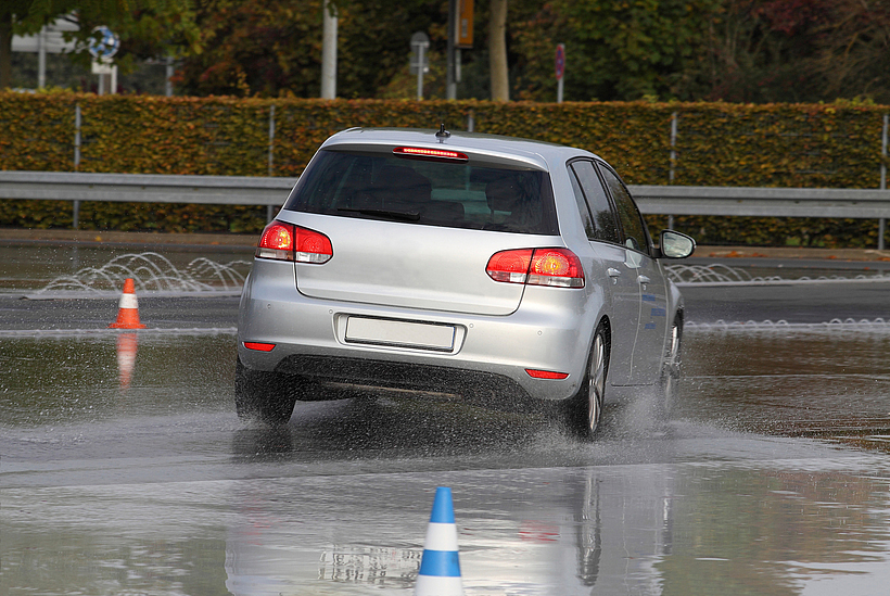 Ein Auto fährt auf einer nassen Straße, auf der zwei Leitkegel aufgestellt sind.