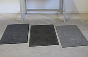 Drei Arbeitsplatzmatten in verschiedenen Grautönen sind auf dem Boden ausgelegt.