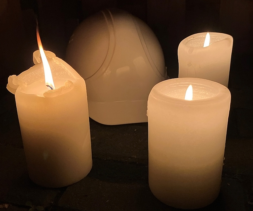 Drei angezündete Kerzen mit einem Schutzhelm in einem abgedunkeltem Raum.