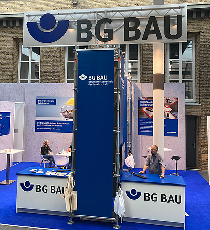 Messestand der BG BAU mit Roll-ups, Tischen und Stühlen.