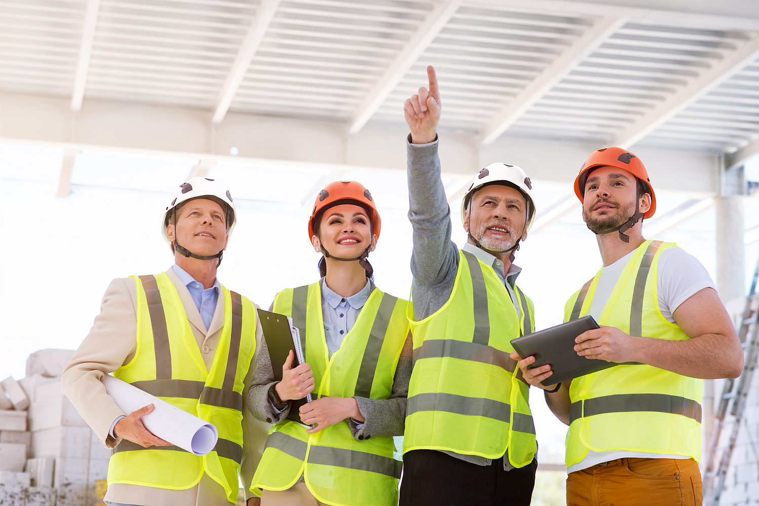 Ein Bauprojektteam bestehend aus drei Männern und einer Frau haben eine Besprechung auf einer Baustelle. Sie tragen alle einen Schutzhelm mit Vier-Punkt-Kinnriemen und Warnwesten.