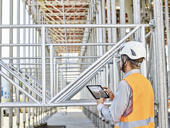 Ein Architekt mit Schutzhelm steht mit einem Tablet in der Hand auf einer Baustelle.