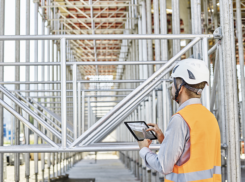 Ein Architekt mit Schutzhelm steht mit einem Tablet in der Hand auf einer Baustelle.