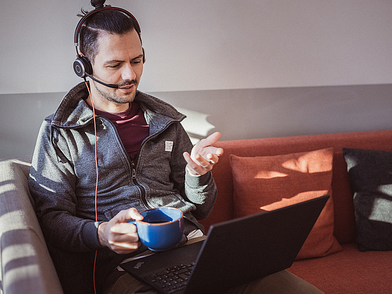 Auf einer Couch sitzt ein junger Mann mit Headset und Laptop auf dem Schoß. Er befindet sich in einer Online-Besprechung.