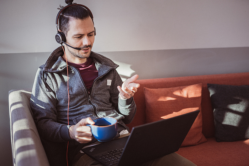 Auf einer Couch sitzt ein junger Mann mit Headset und Laptop auf dem Schoß. Er befindet sich in einer Online-Besprechung.