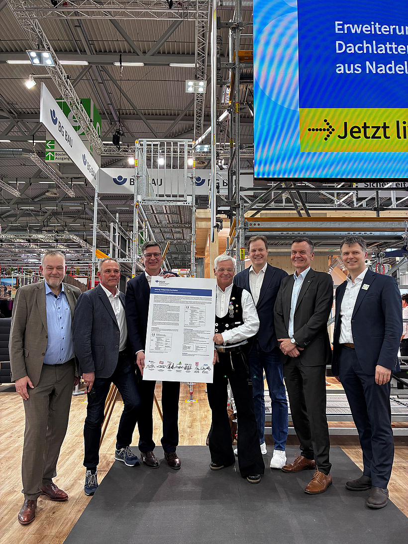 Sieben M&auml;nner stehen auf einem Messestand der BG BAU zusammen und pr&auml;sentieren ein unterzeichnetes Dokument zur Dachlattenvereinbarung vor einer Ger&uuml;stkonstruktion und einer gro&szlig;en Bildschirmanzeige.