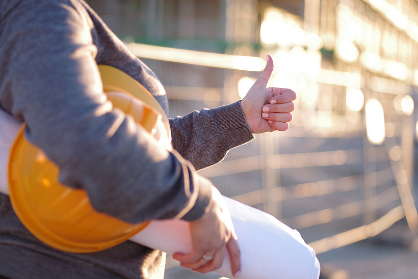 Eine Frau steht mit einem Schutzhelm und Plänen auf einer Baustelle.