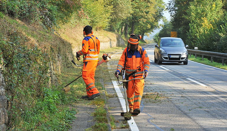 Zwei Männer arbeiten in Warnkleidung an einer Straße. Ein Auto fährt vorbei.