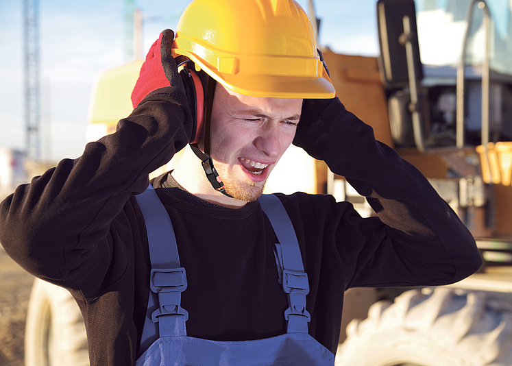 Mann steht auf einer Baustelle und drückt seinen Kapselgehörschutz gegen die Ohren.