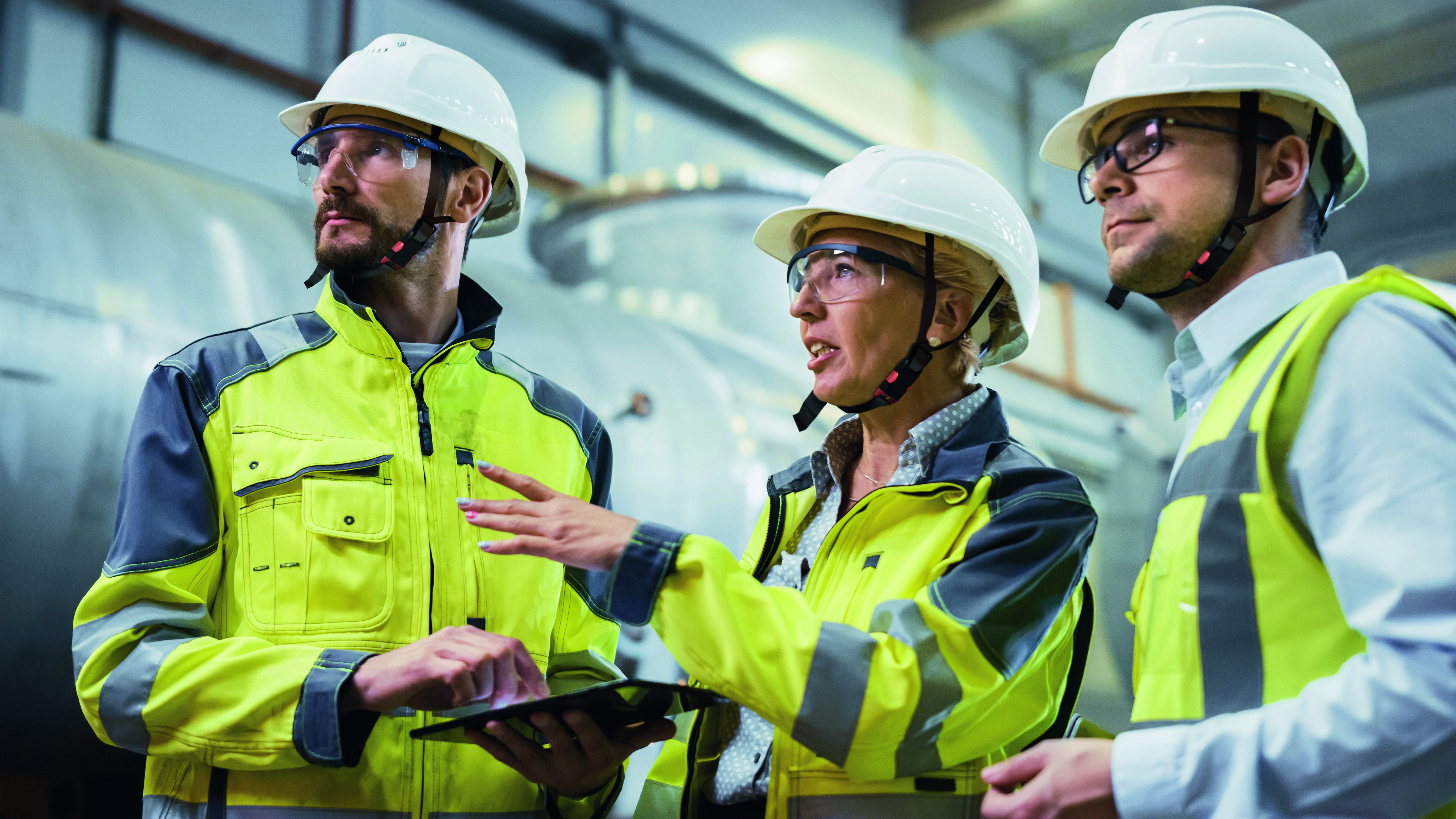 Eine Unternehmerin steht neben zwei Besch&auml;ftigten, von denen einer ein Klemmbrett in der Hand hat, und f&uuml;hrt eine Gef&auml;hrdungsbeurteilung durch. Alle tragen Schutzkleidung.