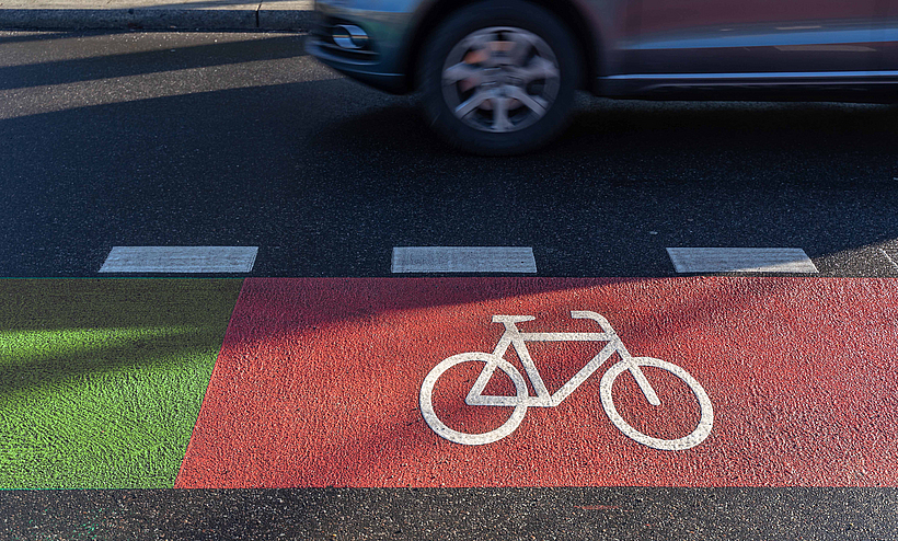 Roter Fahrradweg auf einer Straße. Daneben fährt ein Auto.